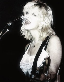 Black and white photo of Courtney Love singing into a microphone on stage with a guitar