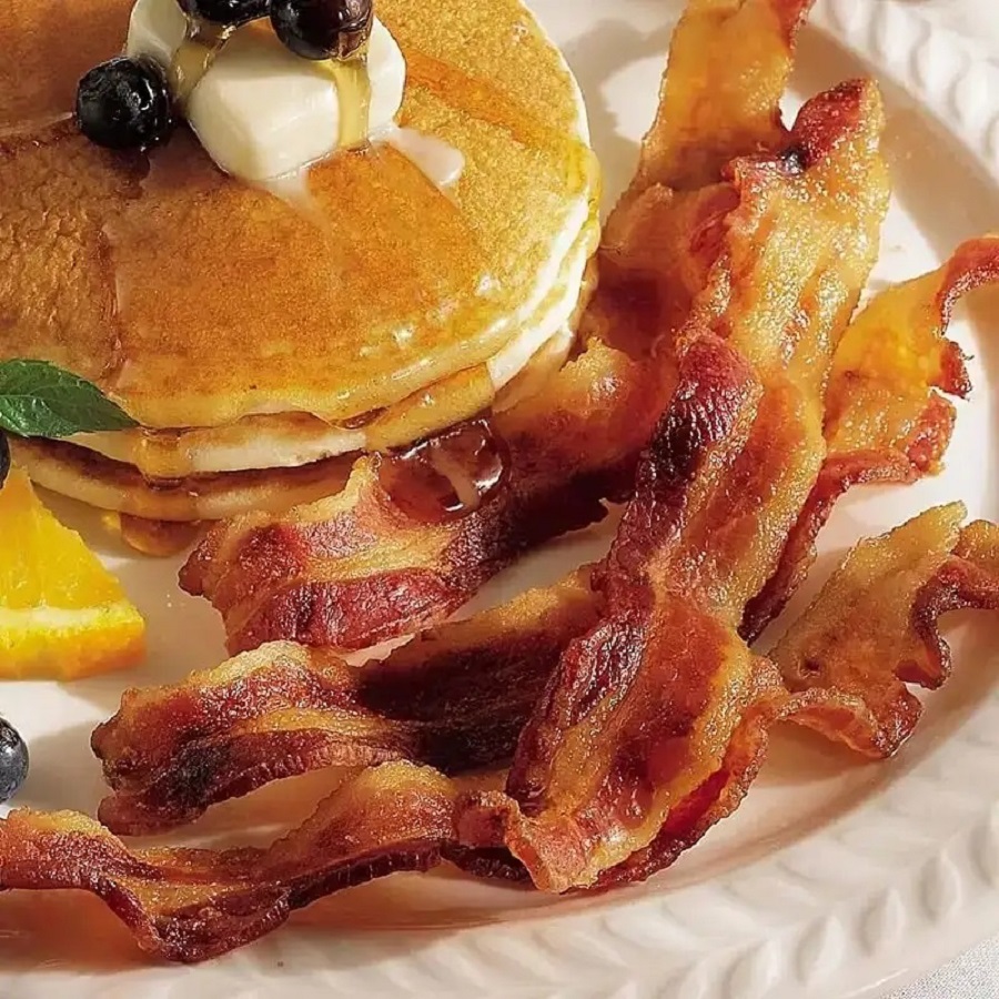 Stack of golden pancakes with butter and blueberries next to crispy bacon strips on a white plate