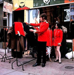 Two people in red coats beside a clothing rack outside a storefront on a sidewalk
