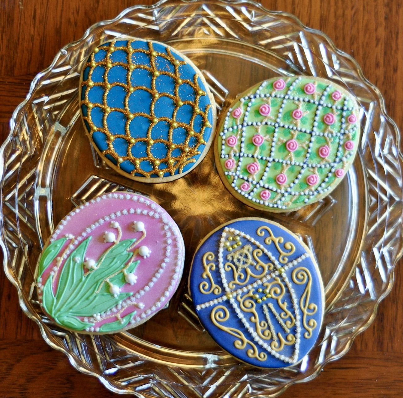 Four decorative Easter egg-shaped cookies with colorful royal icing designs on a glass plate