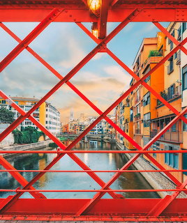 View through red metal bridge lattice showing colorful buildings along a river in Girona, Spain