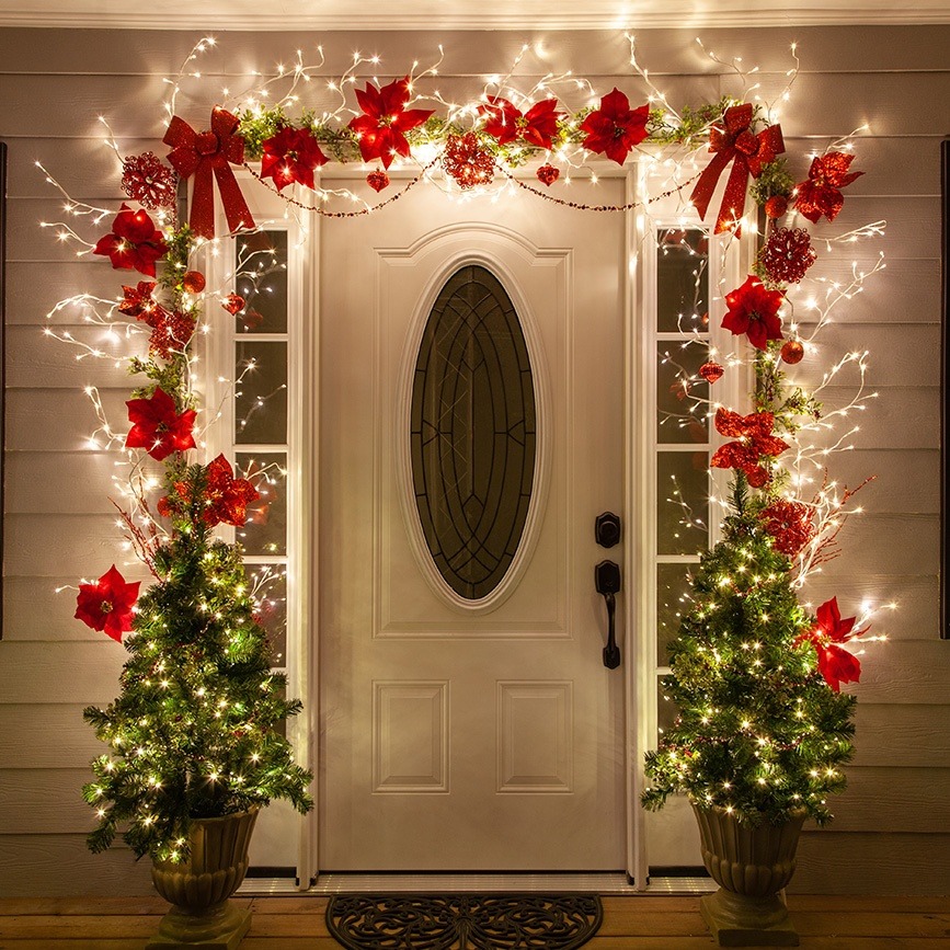Front door decorated with Christmas garland, red flowers, fairy lights, and two small lit Christmas trees