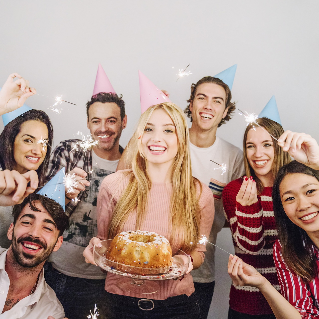 Group of friends in party hats celebrating indoors with sparklers and a bundt cake