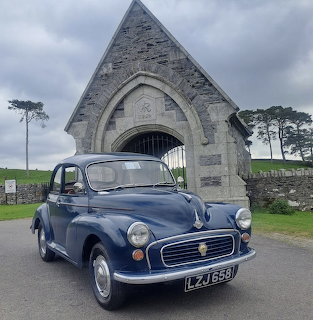 Vintage blue car parked in front of a stone archway on a cloudy day