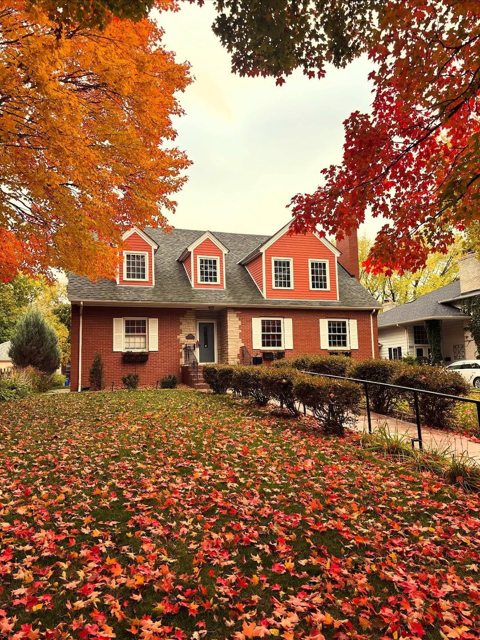Red brick house with dormer windows framed by colorful autumn trees and a carpet of fallen leaves