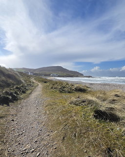 Gravel trail along grassy coastline with beach and hills under cloudy blue sky