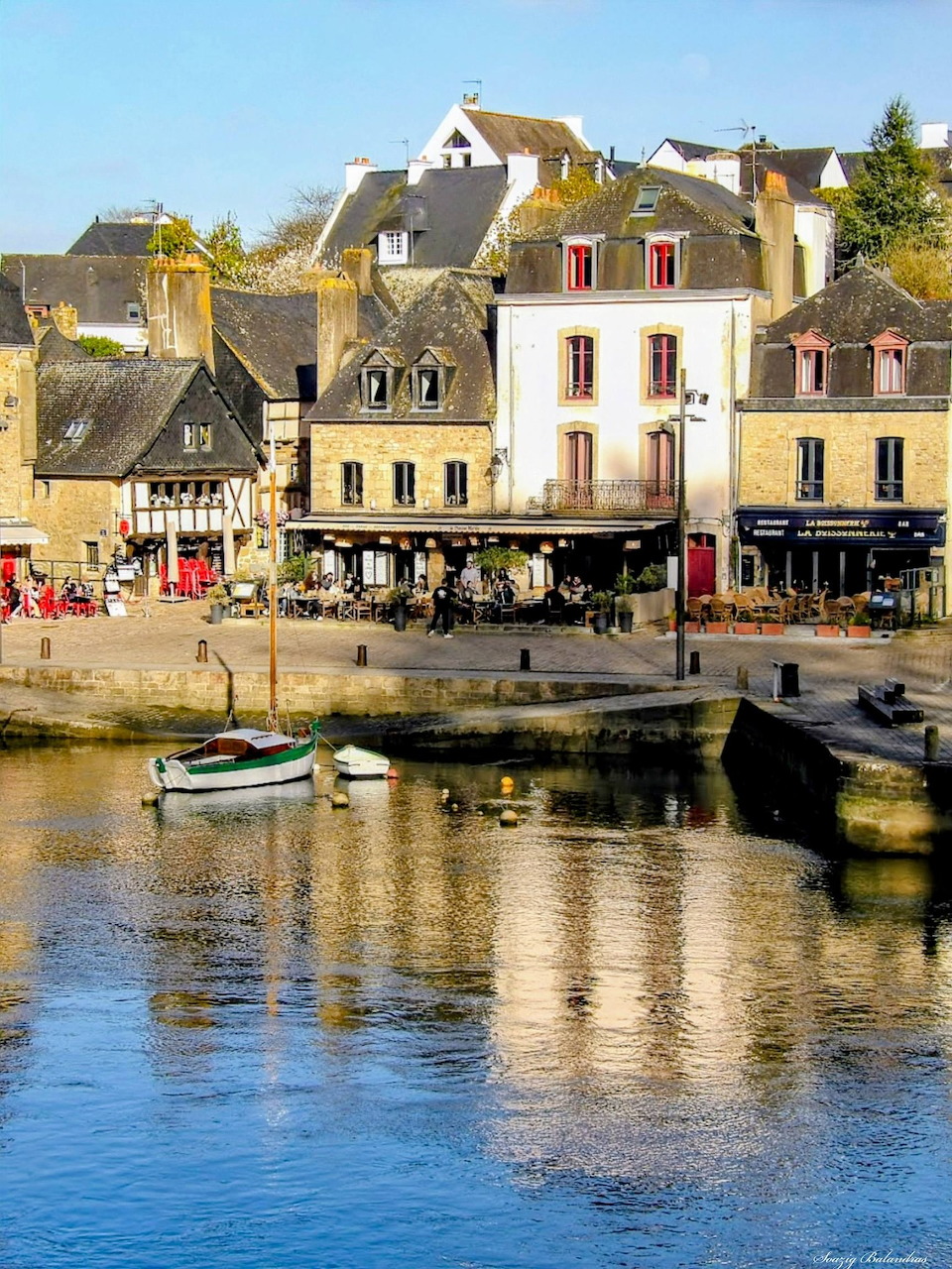 Small boat on calm harbor water reflecting stone buildings and waterfront cafés in a Breton village