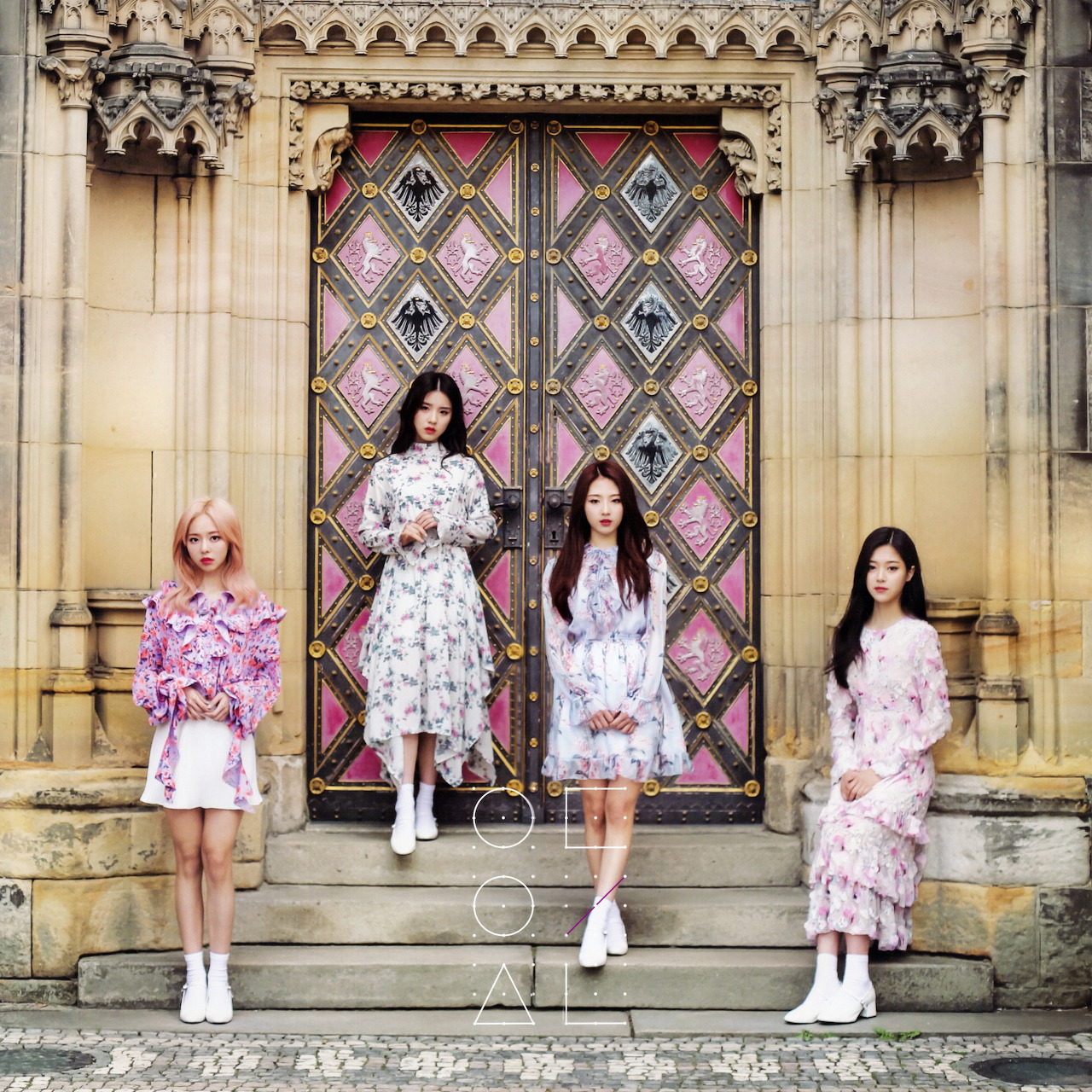 Four young women in pastel dresses standing on stone steps before an ornate arched doorway with pink geometric glass panels