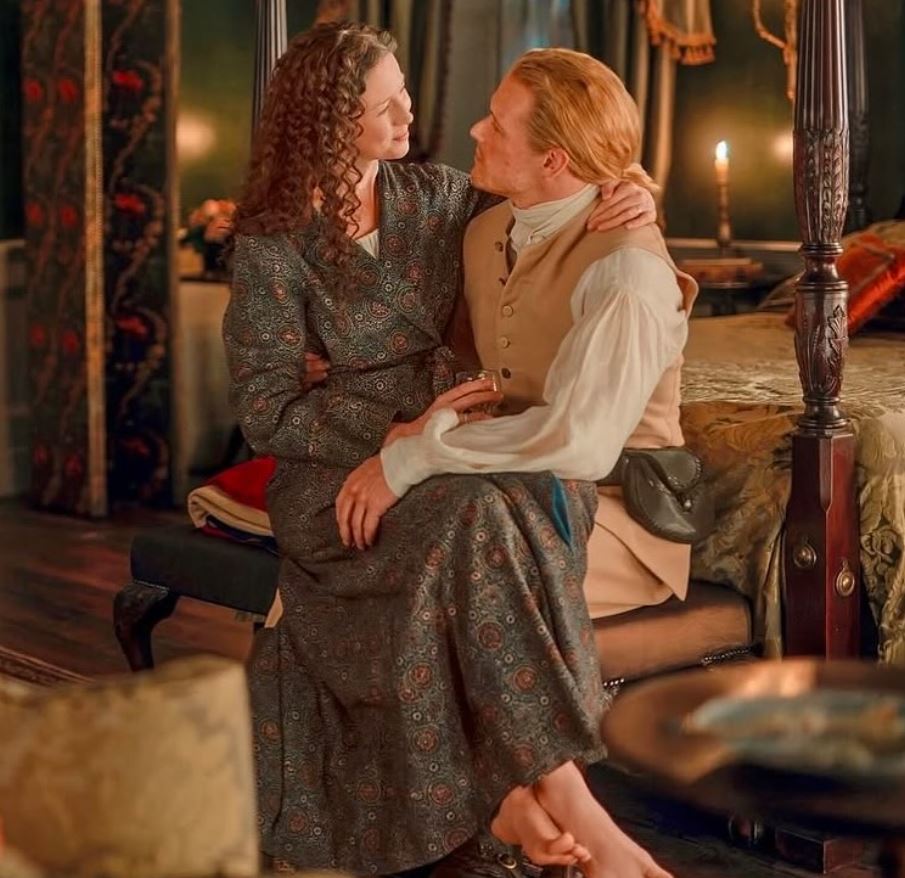 Red-haired couple in historical clothing sitting together on a bed in a candlelit room