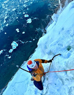 Overhead view of ice climber in orange jacket ascending blue ice wall above water with ice chunks