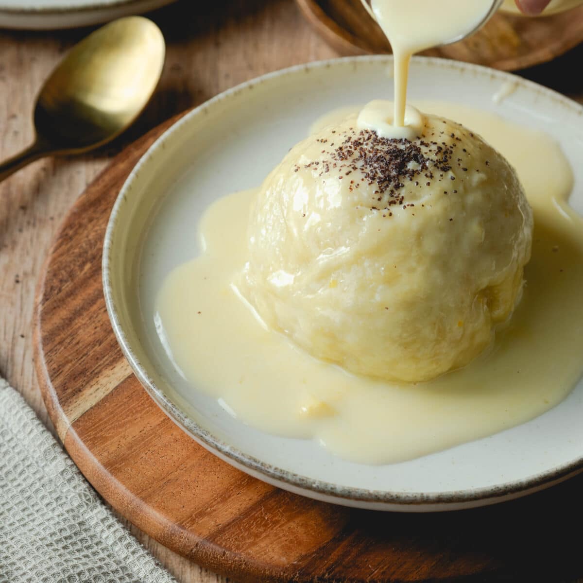Round dumpling on a plate with cream sauce being poured over it, on a wooden cutting board
