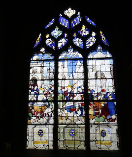 Tall Gothic stained glass window with three panels depicting religious scenes and decorative circular tracery above
