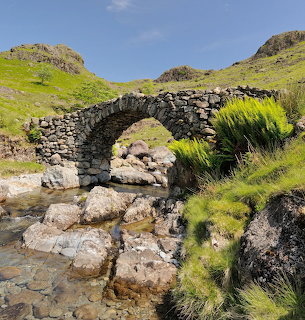 Stone arch bridge spanning a rocky stream in green hills beneath a blue sky