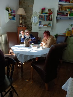 Two women sitting at a round table in a cat cafe, one waving, with bookshelves and warm lighting behind them.
