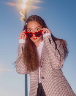 Young woman in a houndstooth blazer wearing red heart-shaped sunglasses against a blue sky