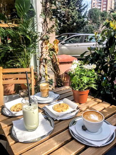 Outdoor cafe wooden table with latte, green drink, cookies, and potted plants on a sunny day.