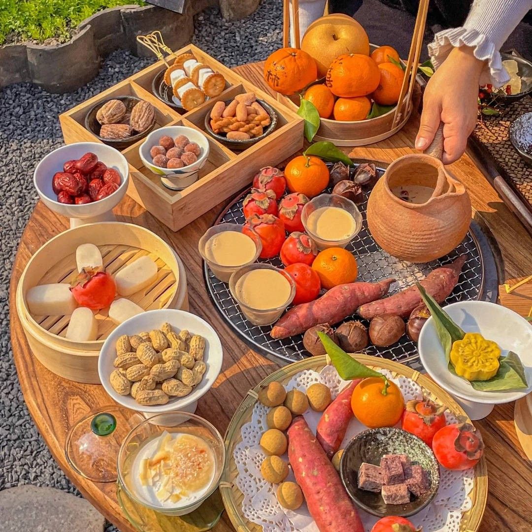 Rustic outdoor table with oranges, tomatoes, nuts, bread, cheese, dips, and sweet potatoes
