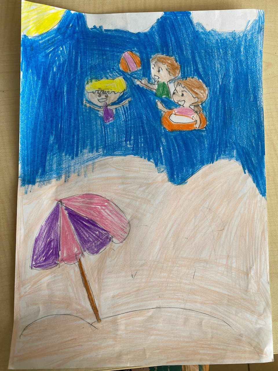 Child's crayon drawing of kids tossing a ball at the beach with a pink umbrella and blue waves