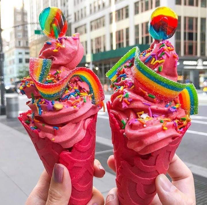 Two pink ice cream cones with rainbow frosting, sprinkles, and lollipops held up in a city street