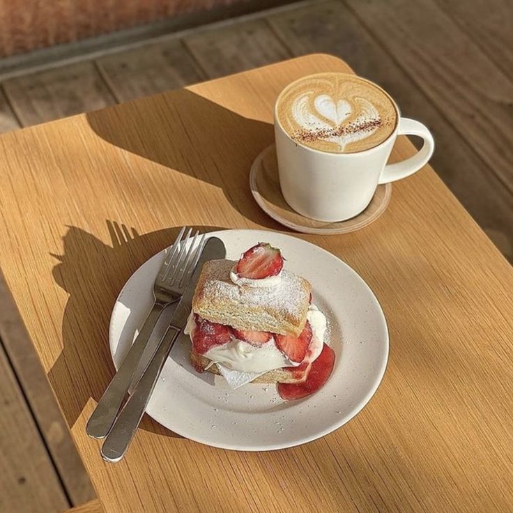 Latte with heart foam art next to a strawberry pastry on a plate atop a wooden table
