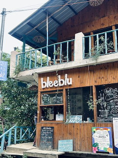 Two-story wooden cafe named Bleeblu with balcony, chalkboard signs, and green plants surrounding it