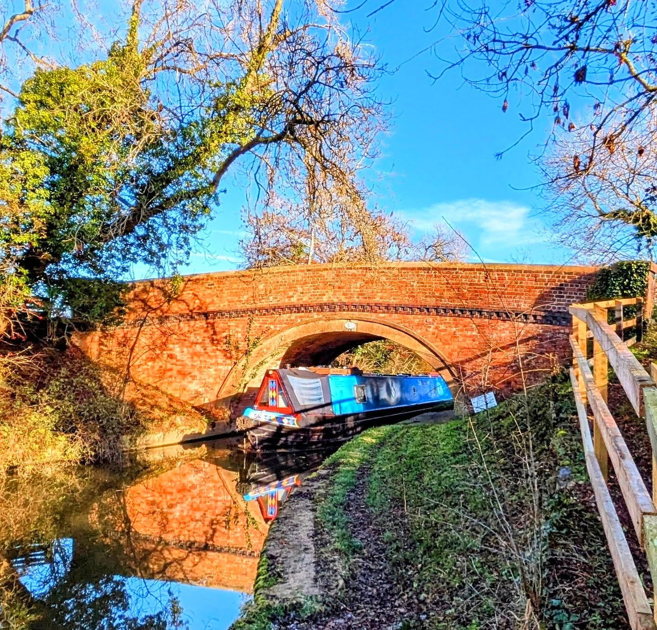 Narrowboat under brick canal bridge with autumn foliage and blue sky reflection