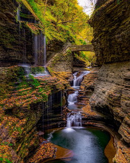 Waterfalls cascading through a narrow gorge with layered rock walls and a stone bridge surrounded by autumn foliage