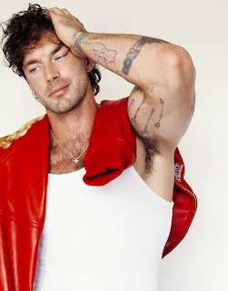 Man with curly hair and arm tattoos in red satin shirt posing with hand in hair
