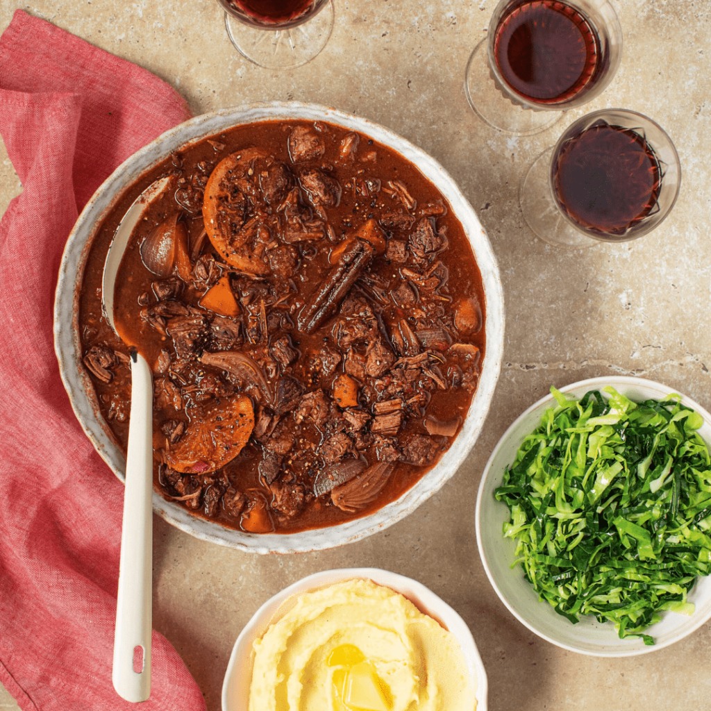 Bowl of beef stew with carrots, accompanied by mashed potatoes and greens