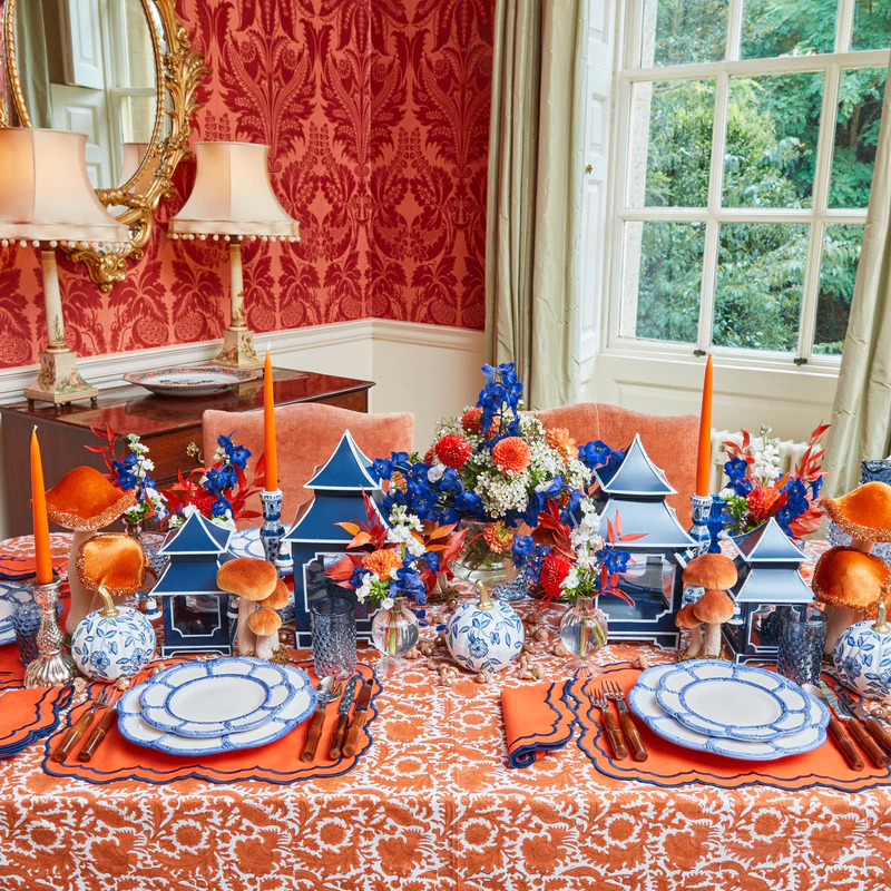 Formal dining table set with orange tablecloth, blue and white china, lamps, and vibrant floral arrangement