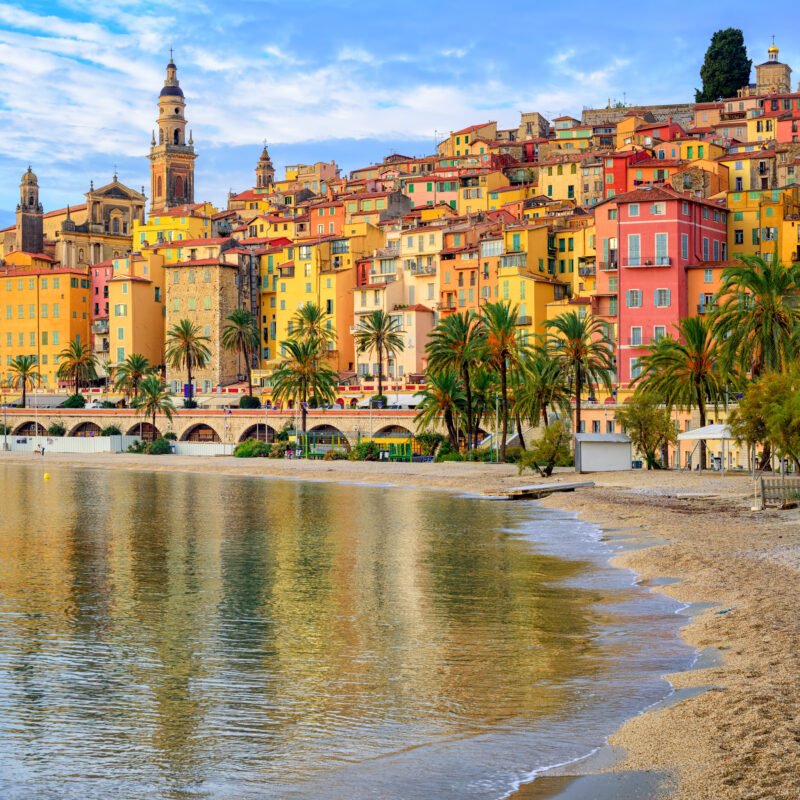 Colorful hillside buildings of Menton, France, with beach and palm trees along Mediterranean coast