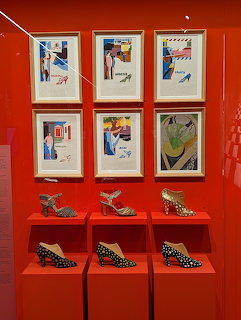 Red museum display case with designer shoes on pedestals and framed fashion illustrations above