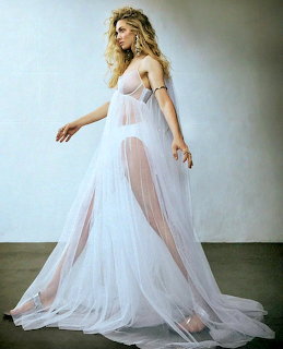 Blonde woman in a flowing sheer white tulle gown posing against a white wall