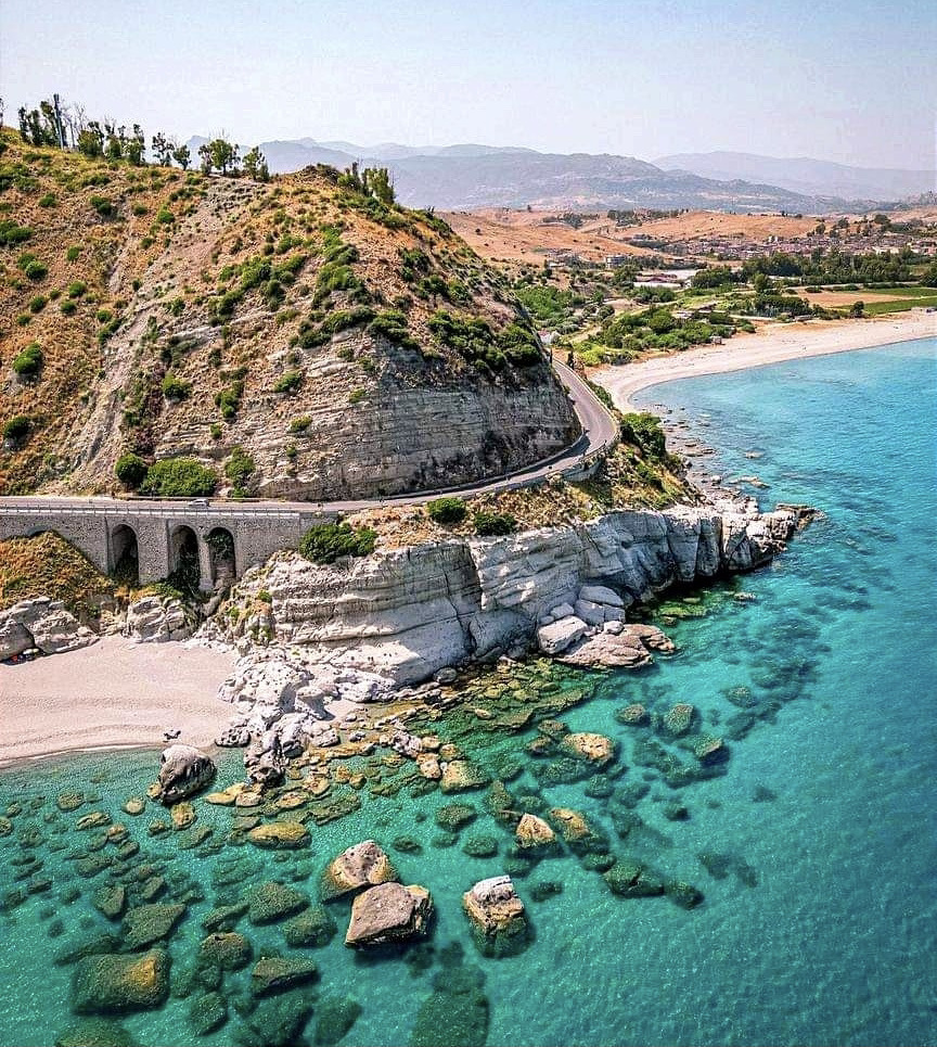 Rocky coastal cliff with a road tunnel, sandy beach, and clear turquoise sea at Capo Bruzzano