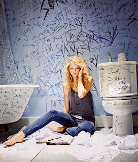 Blonde woman sitting on floor surrounded by toilet paper against wall with handwritten text