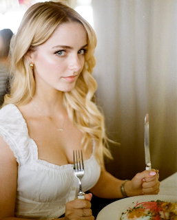 Blonde woman in white dress sitting at table with fork and knife, looking at camera in softly lit indoor setting