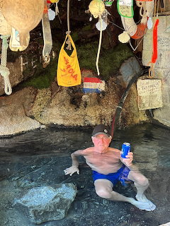 Man sitting in a natural hot spring holding a blue beer can under a rocky overhang with hanging decorations