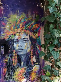 Street art portrait of woman with colorful rainbow feathered headdress on building wall with green ivy
