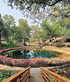 Circular blue spring pool in a landscaped garden with trees, flowers, and wooden railings