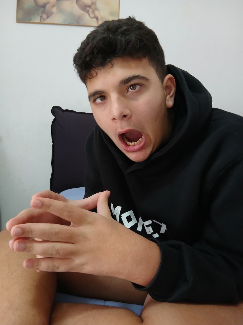 Young man in black hoodie with mouth wide open in shocked expression, sitting indoors