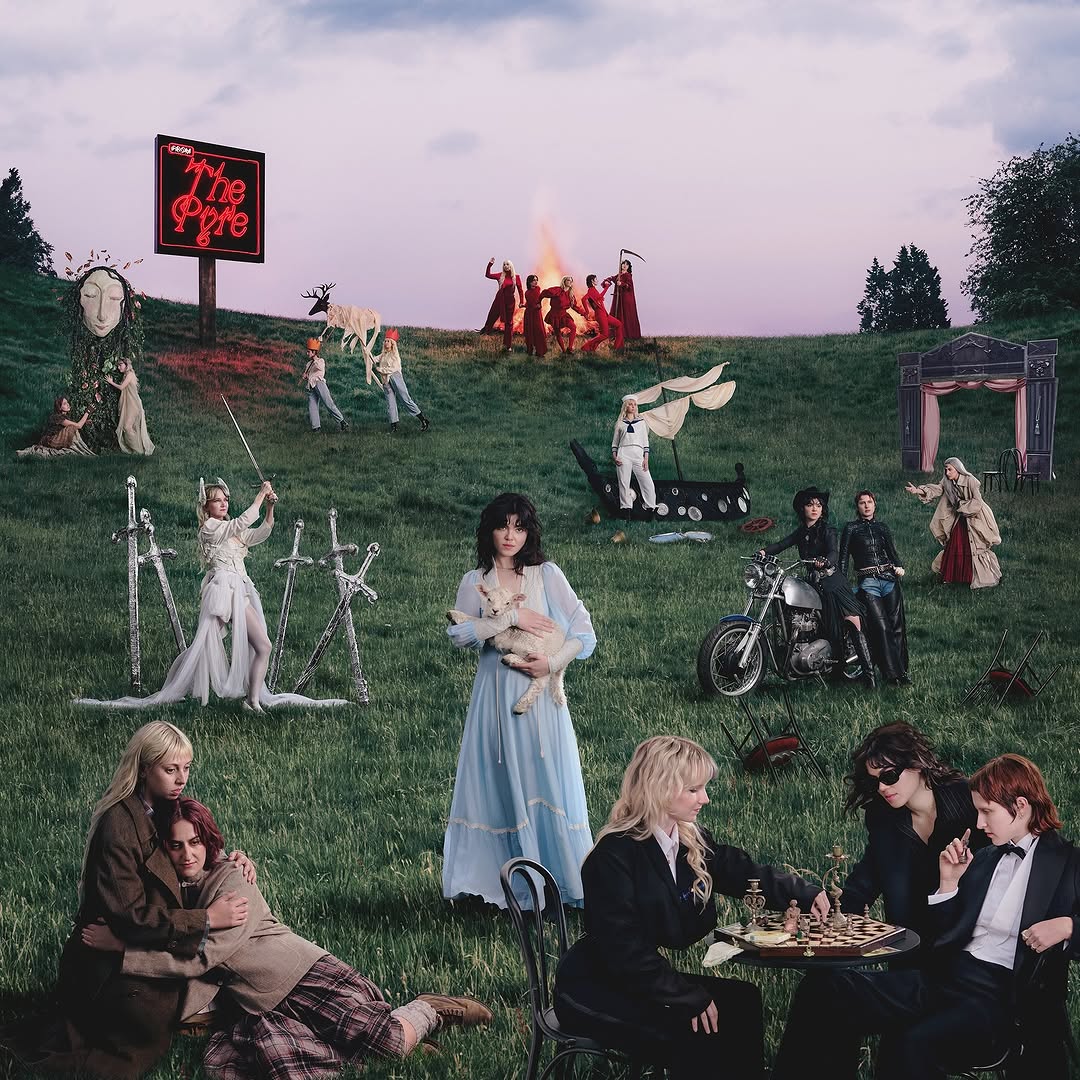 Theatrical outdoor scene with costumed people gathered around a table on grass with a bonfire and neon sign at dusk