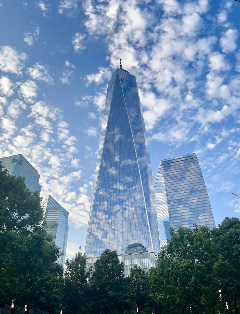 One World Trade Center skyscraper rising into a blue sky with white clouds, trees in foreground