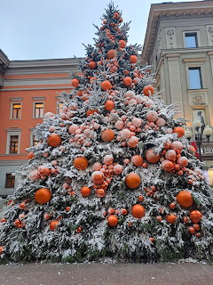 Large snow-covered Christmas tree with orange ornaments in city square with historic buildings