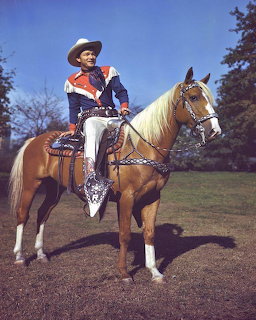 Cowboy in blue and red outfit sitting on palomino horse with white markings outdoors