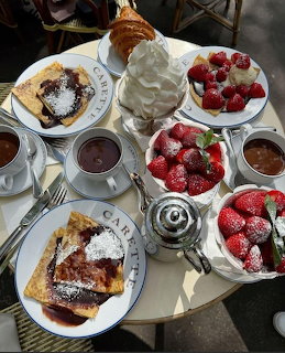 Top-down view of a café brunch table with crepes, fresh strawberries, whipped cream, croissants, and hot chocolate