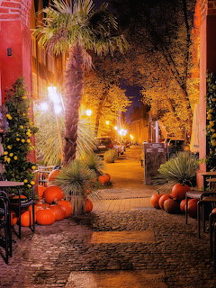 Cobblestone alley at night with pumpkins, glowing streetlights, and autumn trees