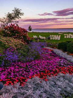 Colorful flower garden with purple and pink blooms overlooking a lake at sunset with white chairs on green lawn