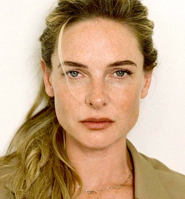 Close-up portrait of a woman with freckles, blue-green eyes, and blonde hair pulled back