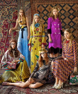 Group of seven women wearing colorful bohemian-style dresses and jewelry posed in decorated room with patterned textiles