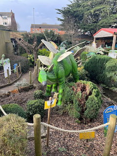 Green dinosaur sculpture in a themed garden surrounded by bushes and pathways